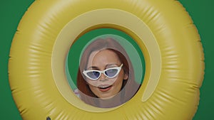 Woman traveller in glasses holds inflatable tube ring and looking through it with amazed face. Isolated on green