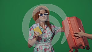 Woman traveller in straw hat and glasses holding suitcase and flight tickets, happy face expression. Isolated on green