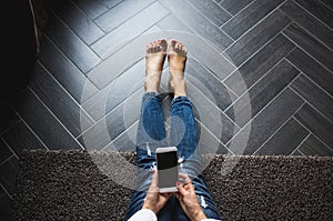 Woman in trendy blue jeans sitting on wooden grey floor, holding blank screen mobile phone. Background, copy space.