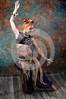 Woman in tribal costume with black lace top and purple pants in red circlet of flowers dancing in front of teal and orange backgro