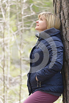Woman at the trunk of a tree. Looks up. Blonde in a blue jacket. Cool weather. Tree bark