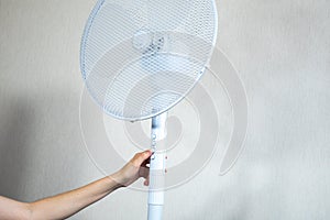 Woman is turning on Metal white modern fan or vent. Working electric floor standing fan in the house. Sunny hot summer day.