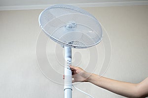 Woman is turning on Metal white modern fan or vent. Working electric floor standing fan in the house. Sunny hot summer day.