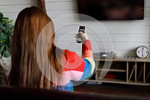 A woman with a TV remote control in her hands in a home interior, view from the back