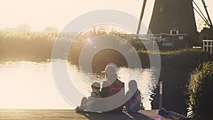 Woman with two kids sit on a lake pier on sunset. Amazing shot of a family together near water edge. 4K back view.