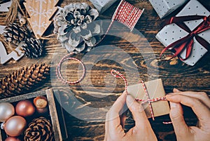 Woman tying gift box wrapped in craft paper with striped white red ribbon. Christmas New Year decoration balls pine cones presents