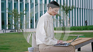 Woman typing laptop sitting on bench near modern building. Lady working remotely