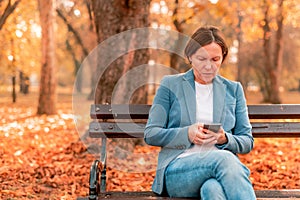 Woman typing text on mobile phone on autumn park bench