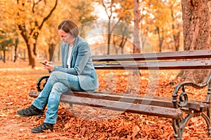 Woman typing text on mobile phone on autumn park bench