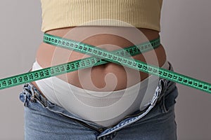 Woman in unfit jeans with measuring tape on light grey background, closeup