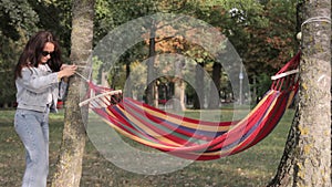 A woman unfolds and ties a hammock to a tree. Outdoor recreation