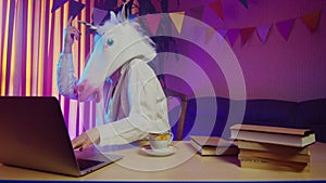 A woman in a unicorn mask is sitting on a chair at a table and typing on a laptop keyboard. This man is happy