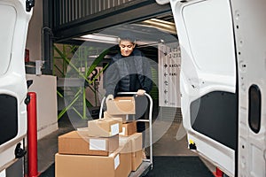 Woman in uniform checking cardboard boxes on a cart before putting them into a van while standing in a warehouse