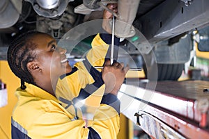 A woman in uniform working underneath a car that is lifted on a hydraulic lift rack, in an automotive repair shop. The mechanic