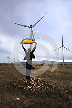 Woman unrecognizable raising a rainbow flag looking towards a cloudy horizon, in a wind farm of wind turbines