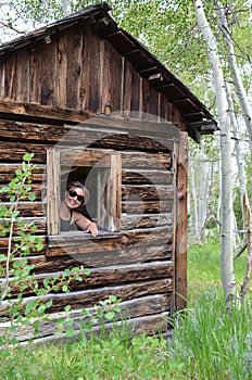 Woman urban explorer smiles through a broken window in an old log cabin in Miners Delight Wyoming