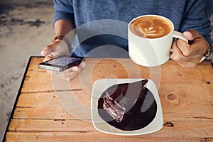 Woman use mobile phone with coffee cup and chocolate cake on wood table in coffee shop.