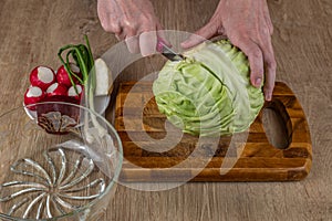 A woman uses a kitchen knife to cut fresh white cabbage on a wooden cutting board. Close-up female hands chopping