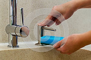 A woman uses a liquid soap dispenser in the kitchen, a sponge for washing dishes. Shiny chrome plated water mixer.