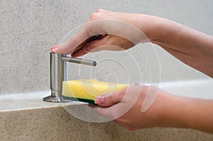 A woman uses a liquid soap dispenser in the kitchen, a sponge for washing dishes. Shiny chrome plated water mixer.