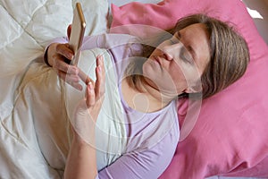 Woman uses mobile phone in a bed. Middle aged 40 years woman lying on pillow and texting on smartphone. Social networking or