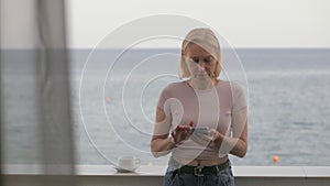 A woman uses a smartphone on the balcony of her hotel room with a sea view.