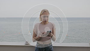 A woman uses a smartphone on the balcony of her hotel room with a sea view.