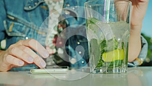 A woman uses a smartphone, next is a glass with a cool cocktail. In the frame are visible only hands. Combine work and