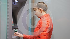 Woman using cash machine. Attractive young female in red bubble jacket inserting credit card into ATM and entering pin