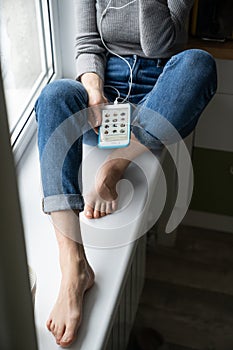 Woman using cellphone, sitting on windowsill listening to podcast or clubhouse - drop-in audio chat.