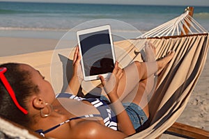 Woman using digital tablet while relaxing on hammock on beach