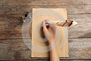 Woman using feather pen to write with ink on parchment at wooden table, top view