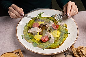 Woman using a fork and a knife to eat a green omelet with a yolk, chicken, cherry tomatoes, greens