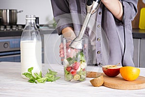 Woman using hand blender to make smoothies. Grey kitchen interior design. Vegetarian, clean eating lifestyle concept