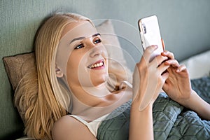 A woman is using her smartphone while she smiles lying on her bed