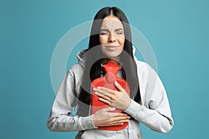 Woman using hot water bottle to relieve pain on light blue background