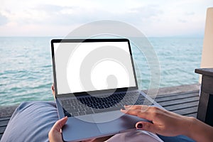 A woman using laptop with blank white desktop screen while sitting by the sea with blue sky background