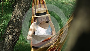 Woman using laptop in hammock. Young female freelancer in hat lying in hammock and working on laptop computer outdoors. Girl openi