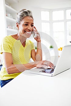 Woman Using Laptop And Talking On Phone In Kitchen At Home