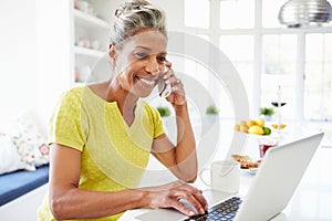 Woman Using Laptop And Talking On Phone In Kitchen At Home