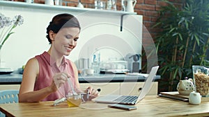 Woman using laptop for video call online. Girl pouring honey into cup of tea.