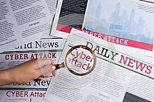 Woman using magnifying glass to read newspaper with headline CYBER ATTACK, top view