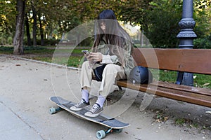 Woman using mobile phone while sitting on a bench in park with her longboard.