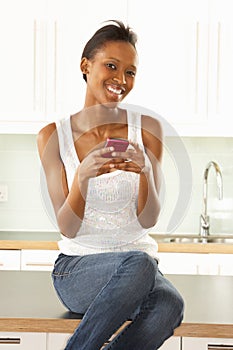 Woman Using Mobile Phone Sitting In Modern Kitchen