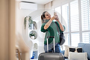 Woman using mobile phone while standing with her suitcase in the hotel room