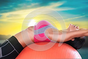 Woman using orange fitness ball to strech her back and exercise her abs on gold tropical sky background