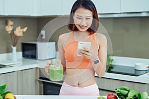 Woman using phone while deinking green smothie and standing in the kitchen