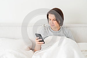 Woman using smartphone as she sits in bed covered with duvet