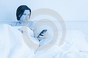 Woman using smartphone as she sits in bed covered with duvet