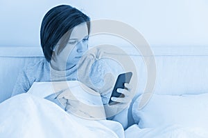 Woman using smartphone as she sits in bed covered with duvet